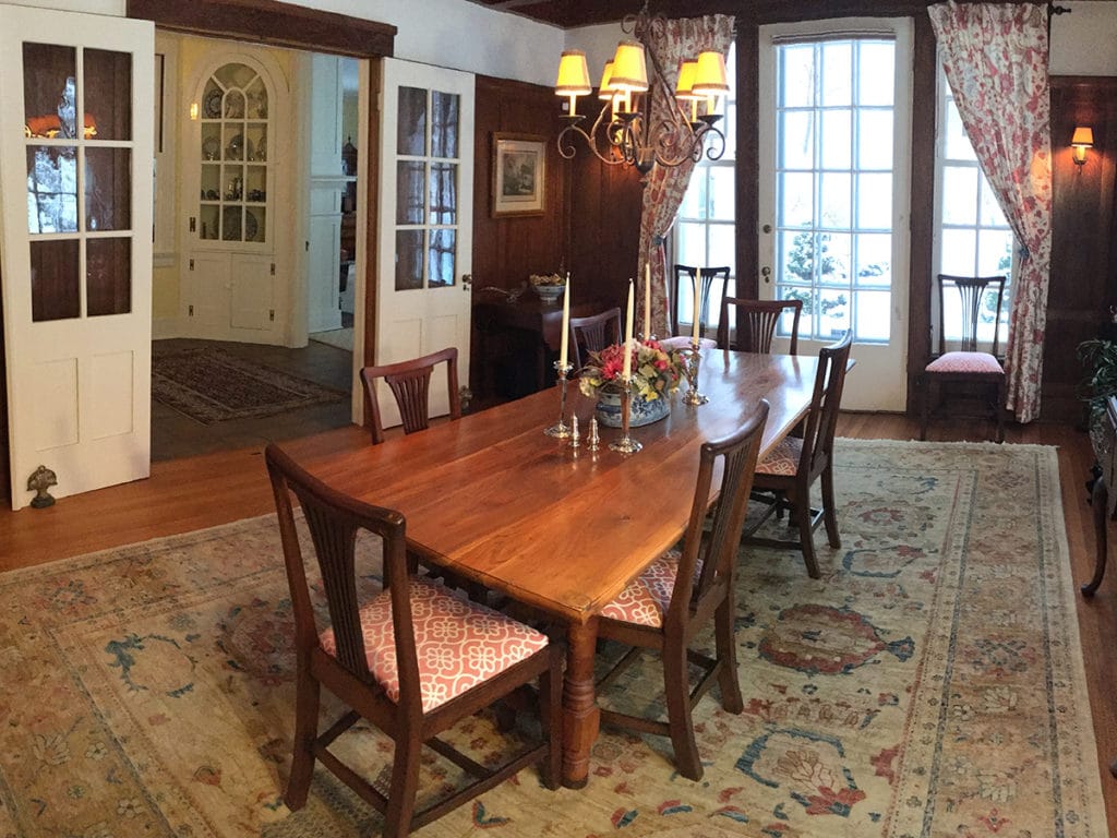 Large hardwood table with 6 chairs on a faded antique rug. Large glass doors and dark wood paneling. the interior door looks out onto a corner cabinet with an arched glass opening