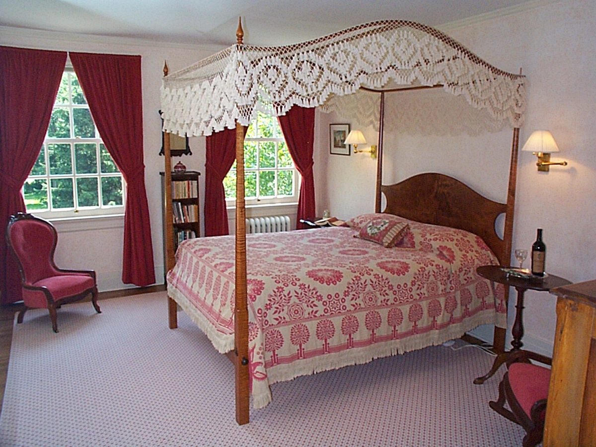 queen size tiger maple canopy bed with white canopy and tassles. Red curtains tied back from 2 large windows. candle-stand with bottle of wine and wine glass