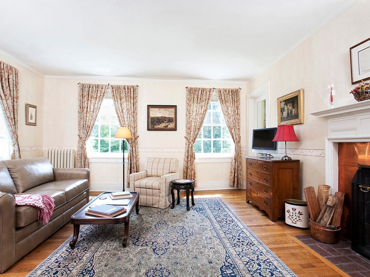 sitting room with leather sofa, coffee table in front. an arm chair with table. an antique 4 drawer chest next to a fireplace with large mantle. wooden floor with large oriental rug in blues and whites.